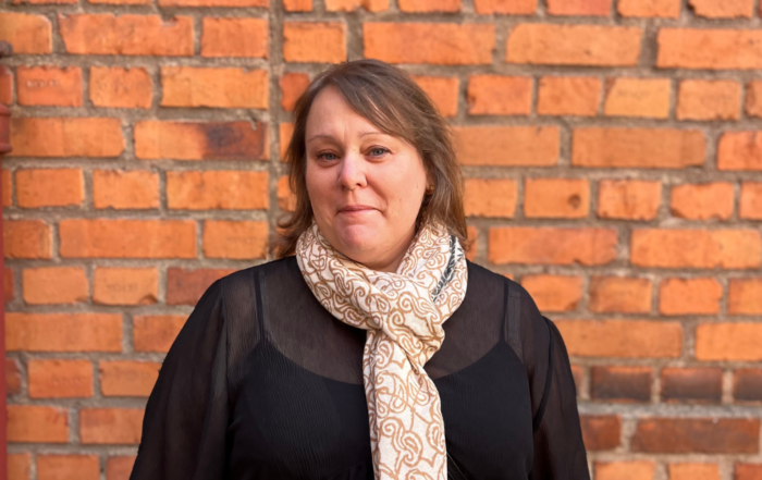 Jenny står framför en tegelvägg och ler mot kameran. Hon är iklädd svarta kläder och en ljus sjal.